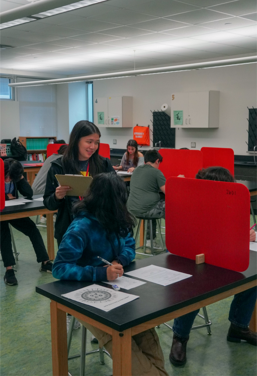 Mariah Vu smiles during seventh period while assisting a student during a test. She saves time by multitasking, carrying around a clipboard, grading papers and proctoring the test.