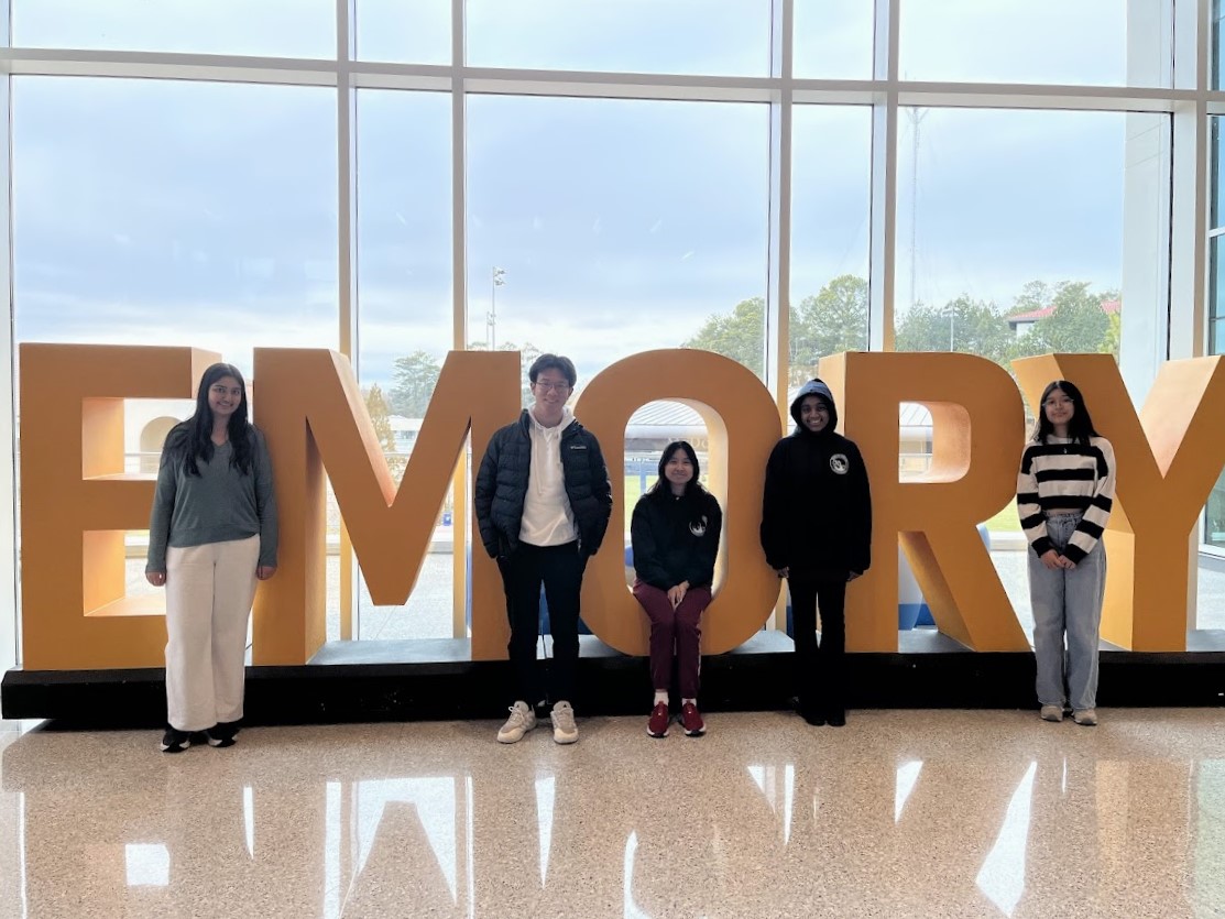 Congressional debaters Alia Hassan, Andy Shen, Samantha Tran, Simone Goffney and Catherine Xue smile in front of the yellow Emory sign in the Northern Emory Student Center. They spectated the final round of Congressional Debate after Shen, Tran and Xue were eliminated in semi-finals.