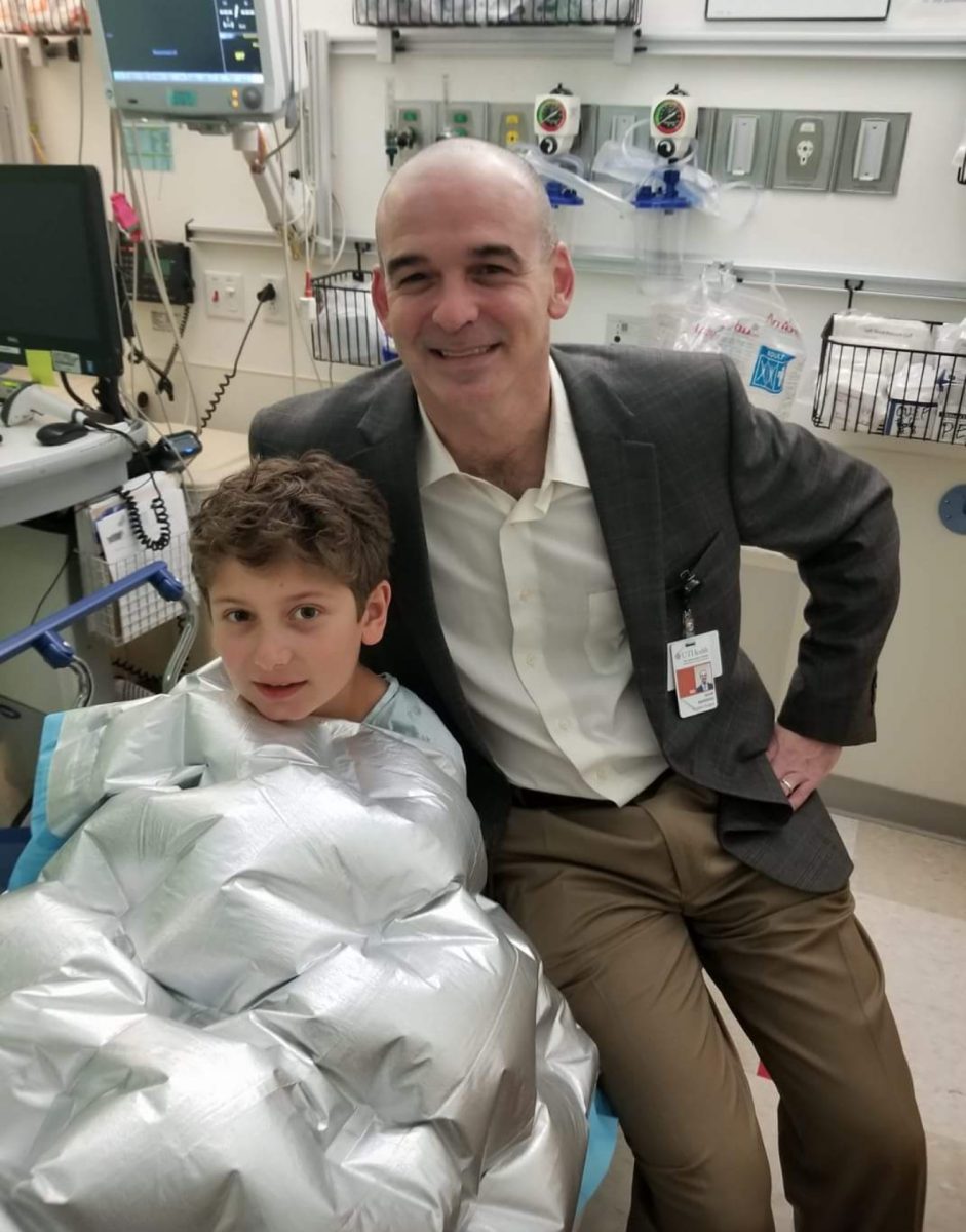Eight-year-old Adam Kay sits next to his pediatric neurosurgeon, Dr. Sandberg. Kay is wrapped in a hospital blanket to keep himself warm.
