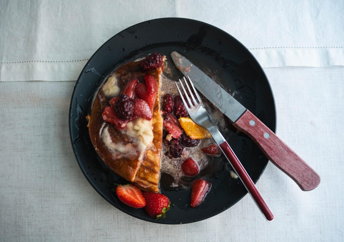 A half-eaten plate of the maple honey butter pancakes covered in a fruit compote sits on a white tablecloth. The berry compote took one hour to make after cooking down sugar, strawberries, blackberries, orange slices and added zest.